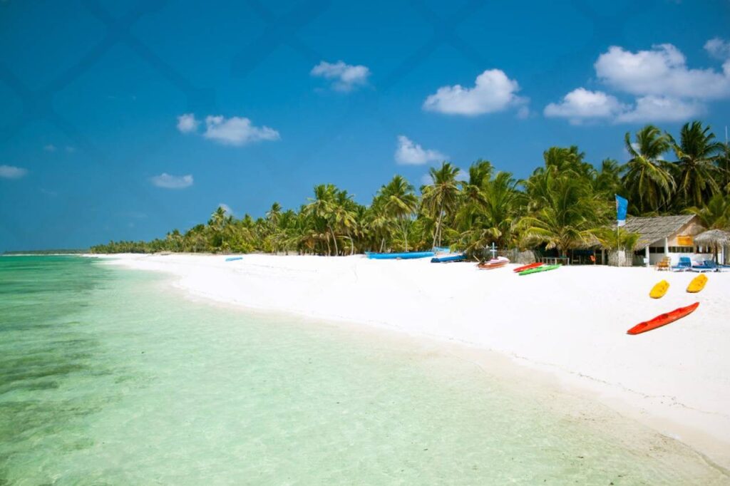 A scenic beach on Agatti Island featuring palm trees and a few boats anchored in the clear blue water.