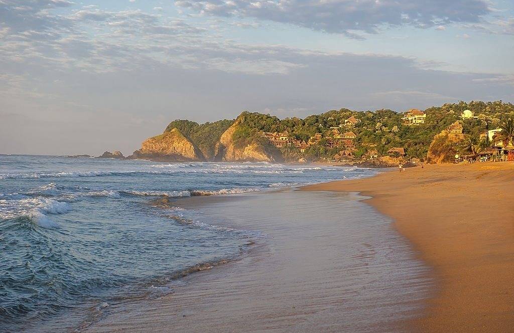 A scenic view of Playa Zipolite beach with gentle waves and a lush hillside in the background.