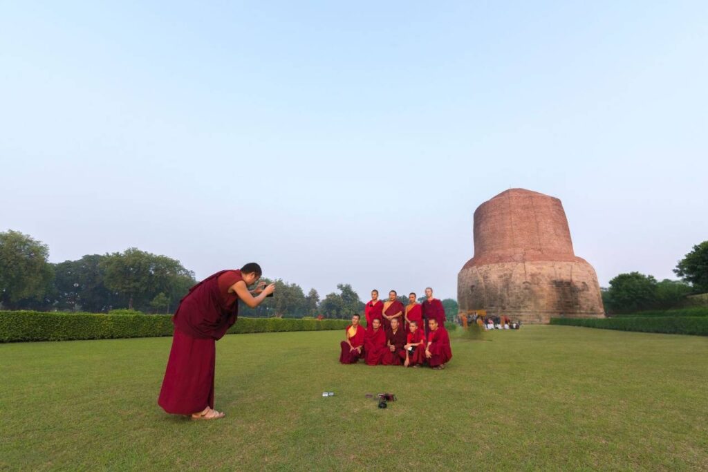 A group of monks in red robes poses for a photo at Sarnath, surrounded by a serene landscape.