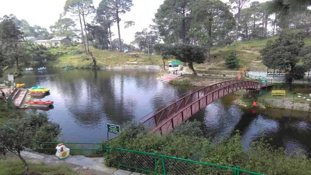 A park bridge crosses a tranquil Bhulla Lake, Lansdowne, surrounded by greenery, with boats leisurely drifting nearby.