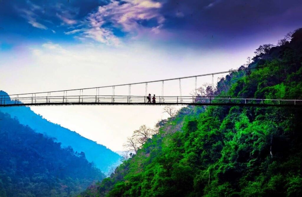 A couple walks hand-in-hand across a suspension bridge over the Dawki River, surrounded by lush greenery.