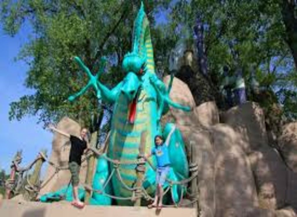 Two kids take a picture in front of a massive happy green dragon statue at an outdoor amusement park.